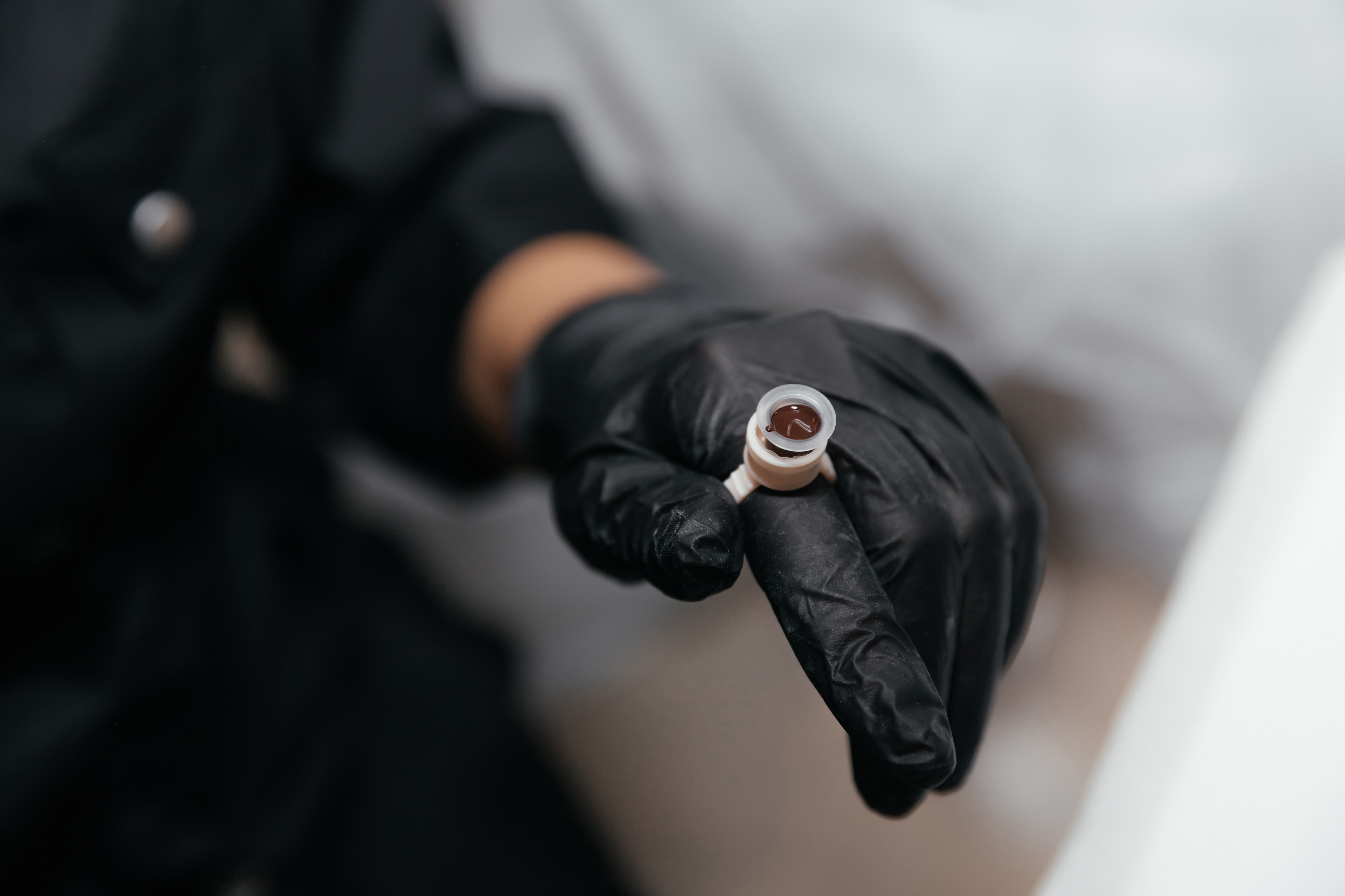 Finger in a black glove is wearing an permanent paint ring with brown pigment. Close-up of pigment container for eyebrow or lip tattoo. Beauty procedure preparation. Selective focus, close-up